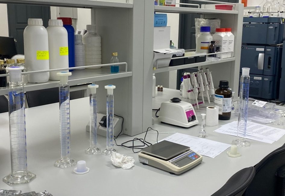 A bench in Kimagri's OECD-GLP certified laboratory: graduated cylinders, a precision balance, pipettes and reagent bottles, with analytical instruments behind.