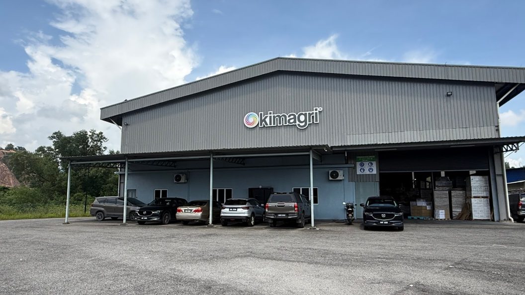 The Kimagri factory at Kampung Kepayang: a grey steel-clad building carrying the Kimagri sign, with limestone hills behind it.